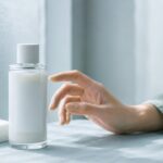 乳白色の化粧水ボトルとコットンに手を伸ばして迷う女性の手元