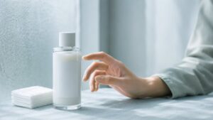 乳白色の化粧水ボトルとコットンに手を伸ばして迷う女性の手元